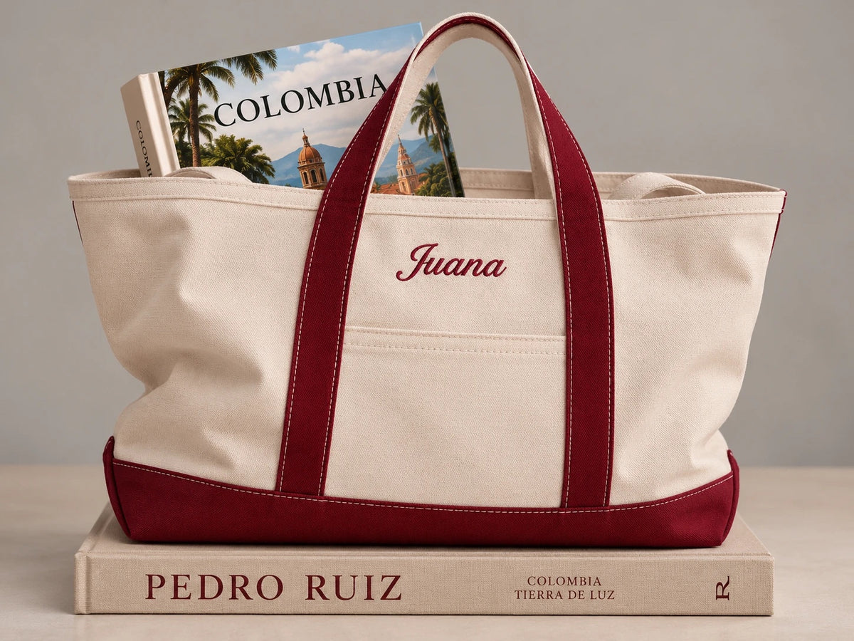 Tote natural con base y asas vino, con el nombre Juana bordado, sobre un libro de Colombia